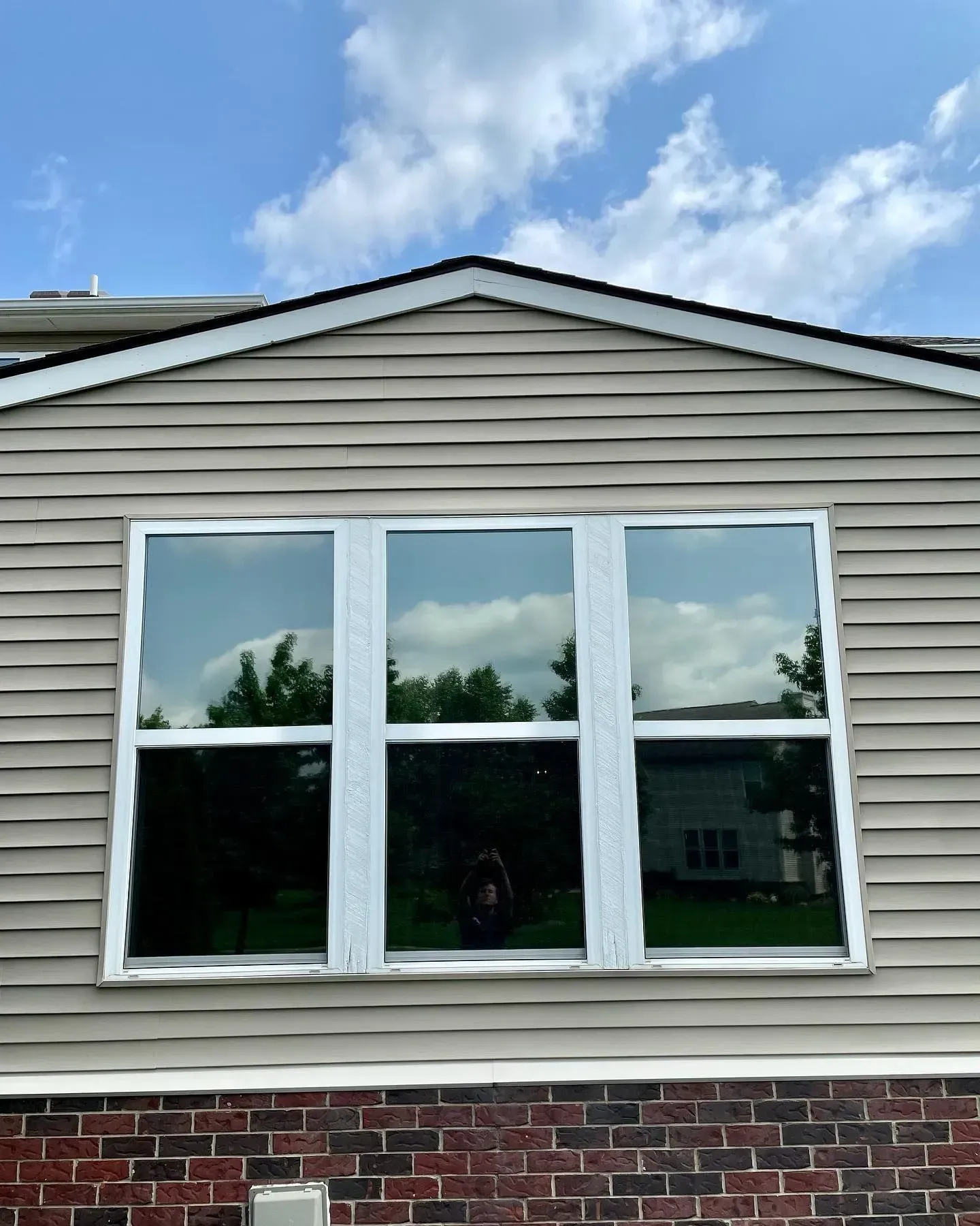 Three-pane white window on a beige house above a red brick base under a blue sky
