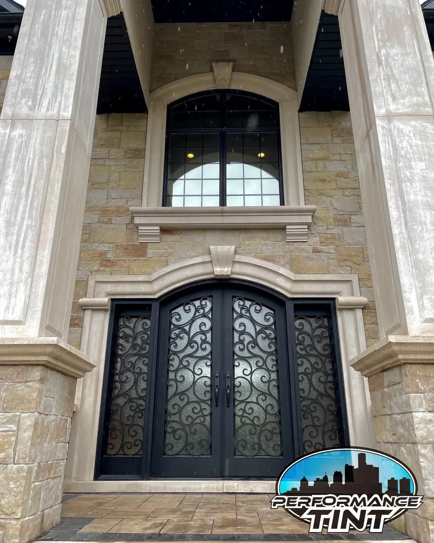 Elegant stone house entry with black wrought-iron double doors and arched window above.