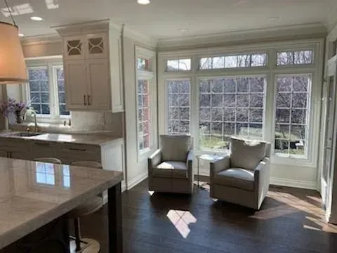 Bright kitchen and sunroom with two gray chairs by large windows and a marble island