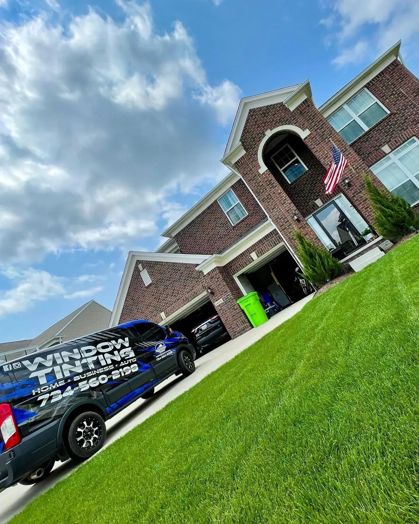 Black service van parked beside a brick house with an American flag and bright green lawn under a blue sky