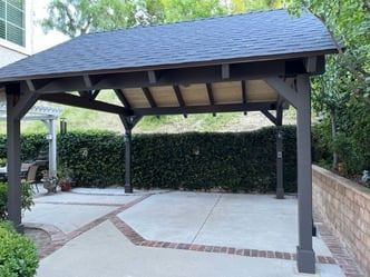 A large wooden gazebo with a slate roof is sitting in the middle of a driveway.