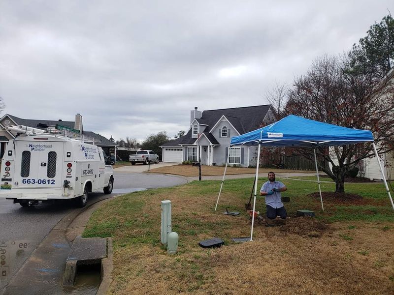 A worker kneels under a blue canopy on a cloudy day, near a utility truck and suburban homes.