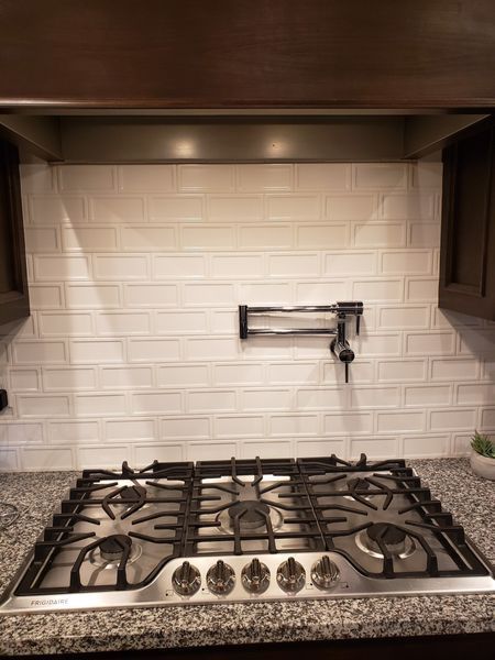 Gas stovetop with a pot filler against a white brick-style tiled backsplash, granite countertop, and dark cabinets.