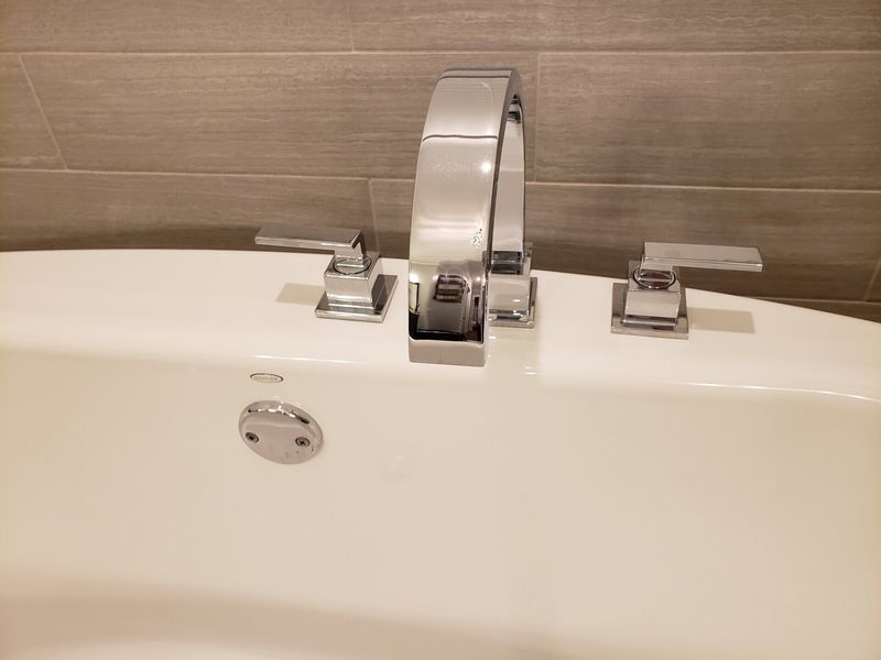 Close-up of a chrome bathtub faucet and handles on a white tub, with a gray tile background.