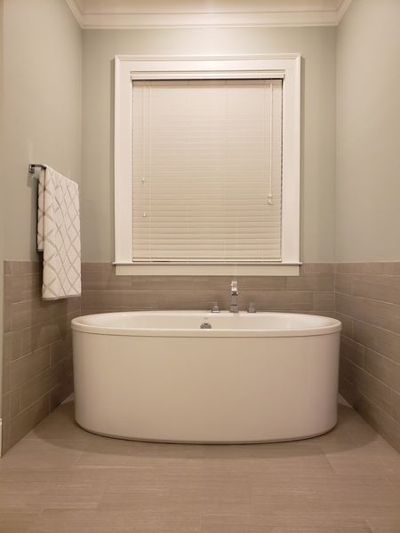 Oval bathtub under a window with closed blinds, in a bathroom with pale gray walls and neutral tile.