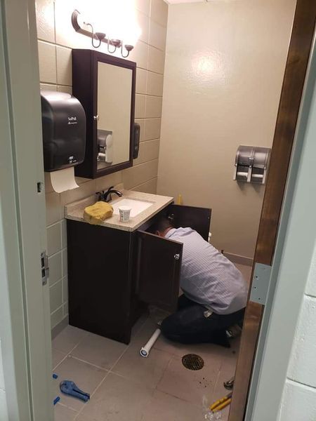 Man kneeling, working on plumbing under a bathroom sink. Beige walls, tile floor, sink and mirror visible.