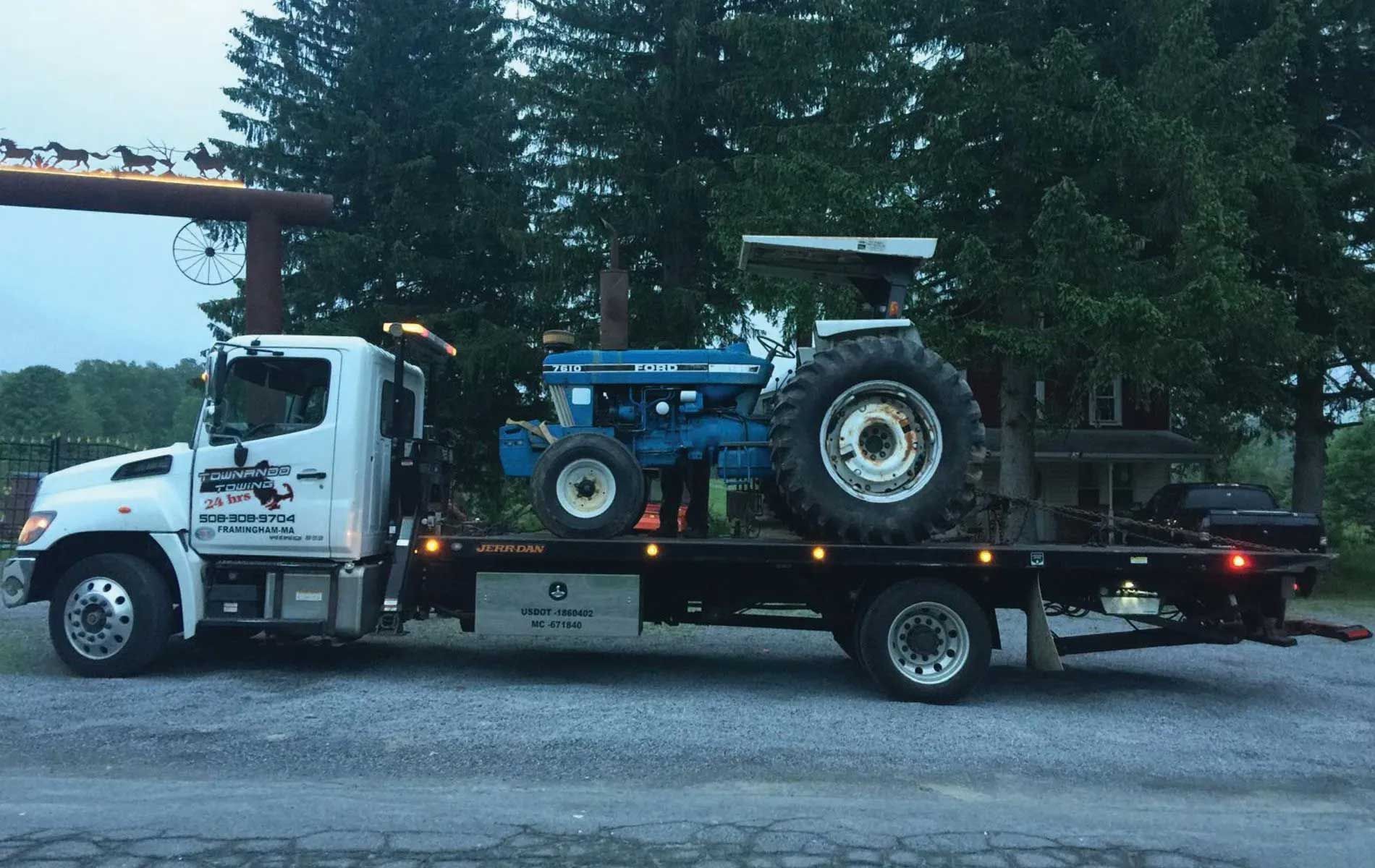 White tow truck hauling a blue tractor at dusk. Trees and a decorative arch in the background.