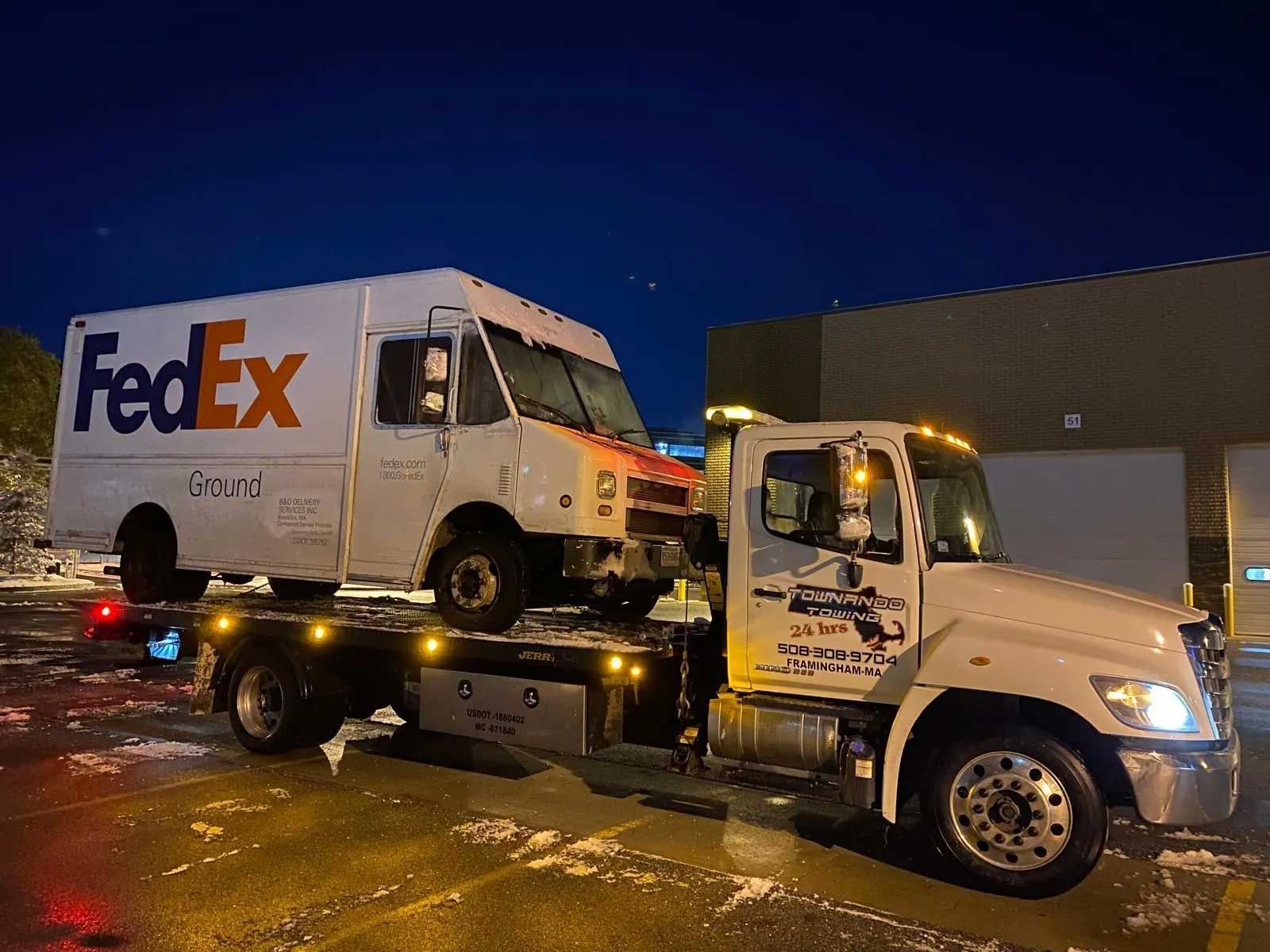 Tow truck carrying a damaged FedEx delivery truck at night.