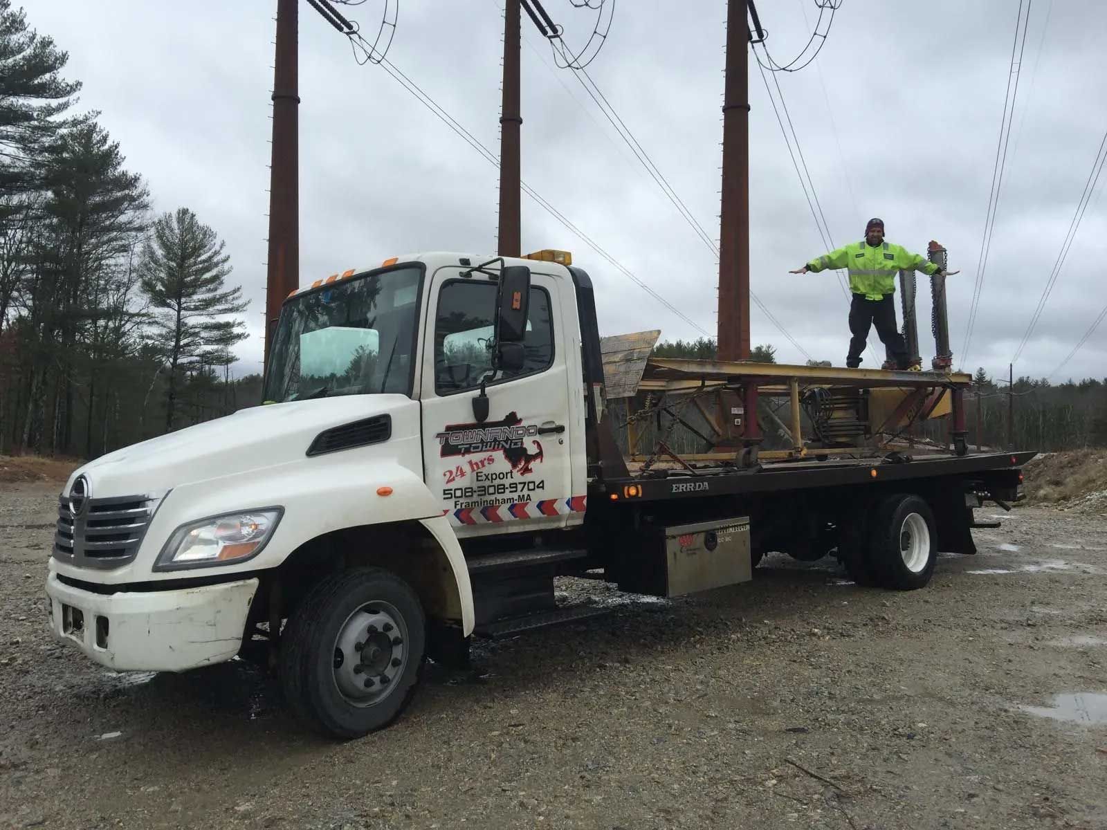 White truck with a worker on the flatbed, near power lines. Worker in neon vest, arms outstretched.