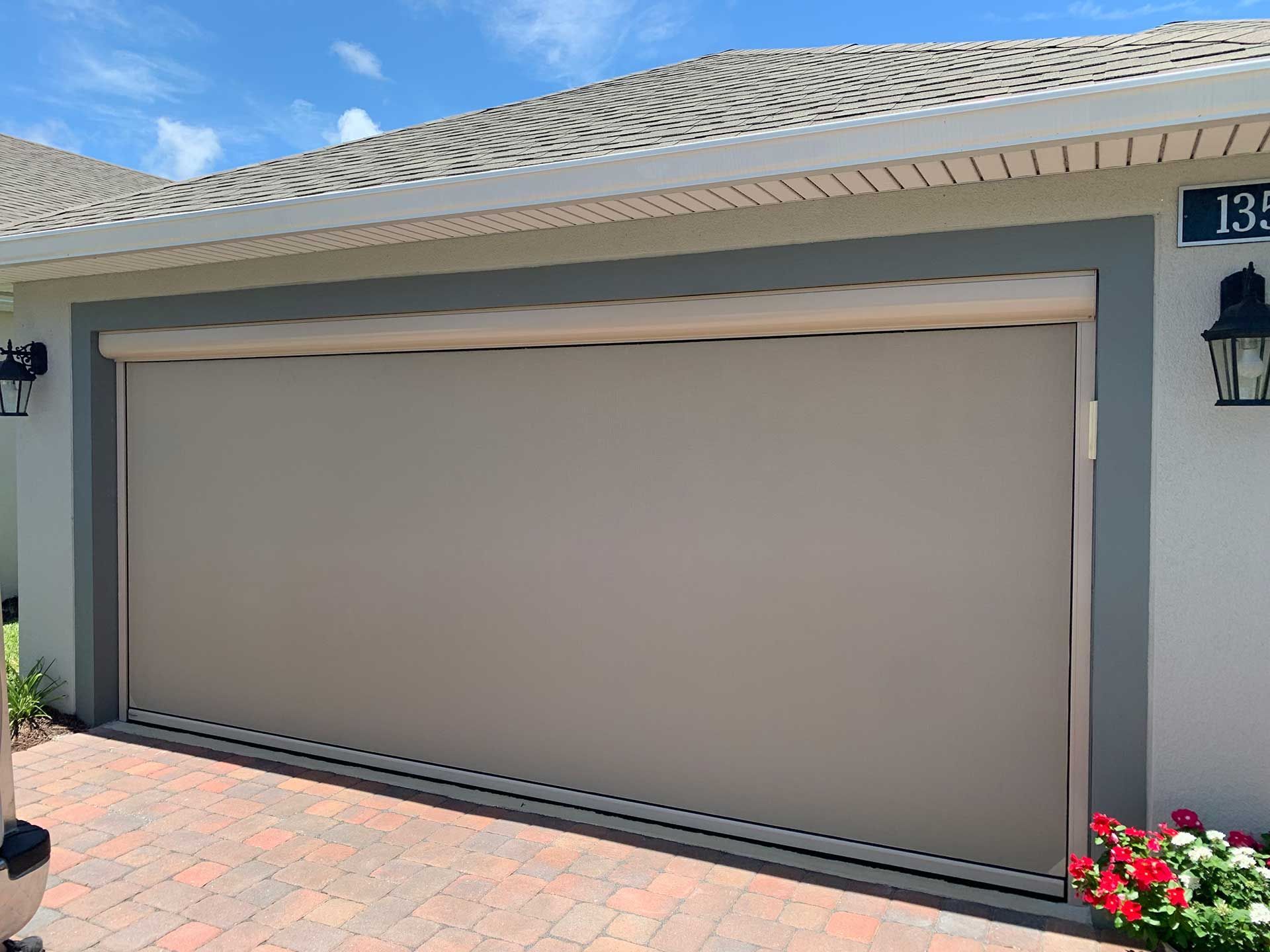 Closed beige rolling garage screen on a stucco house with tiled roof and flowering plants by the entrance