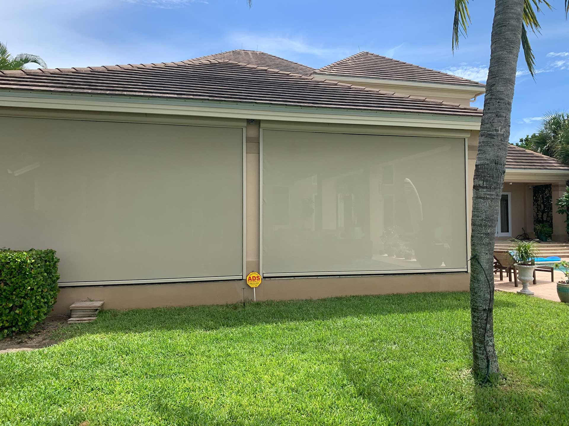 House with closed beige shutters, green lawn, and a palm tree in a sunny backyard.