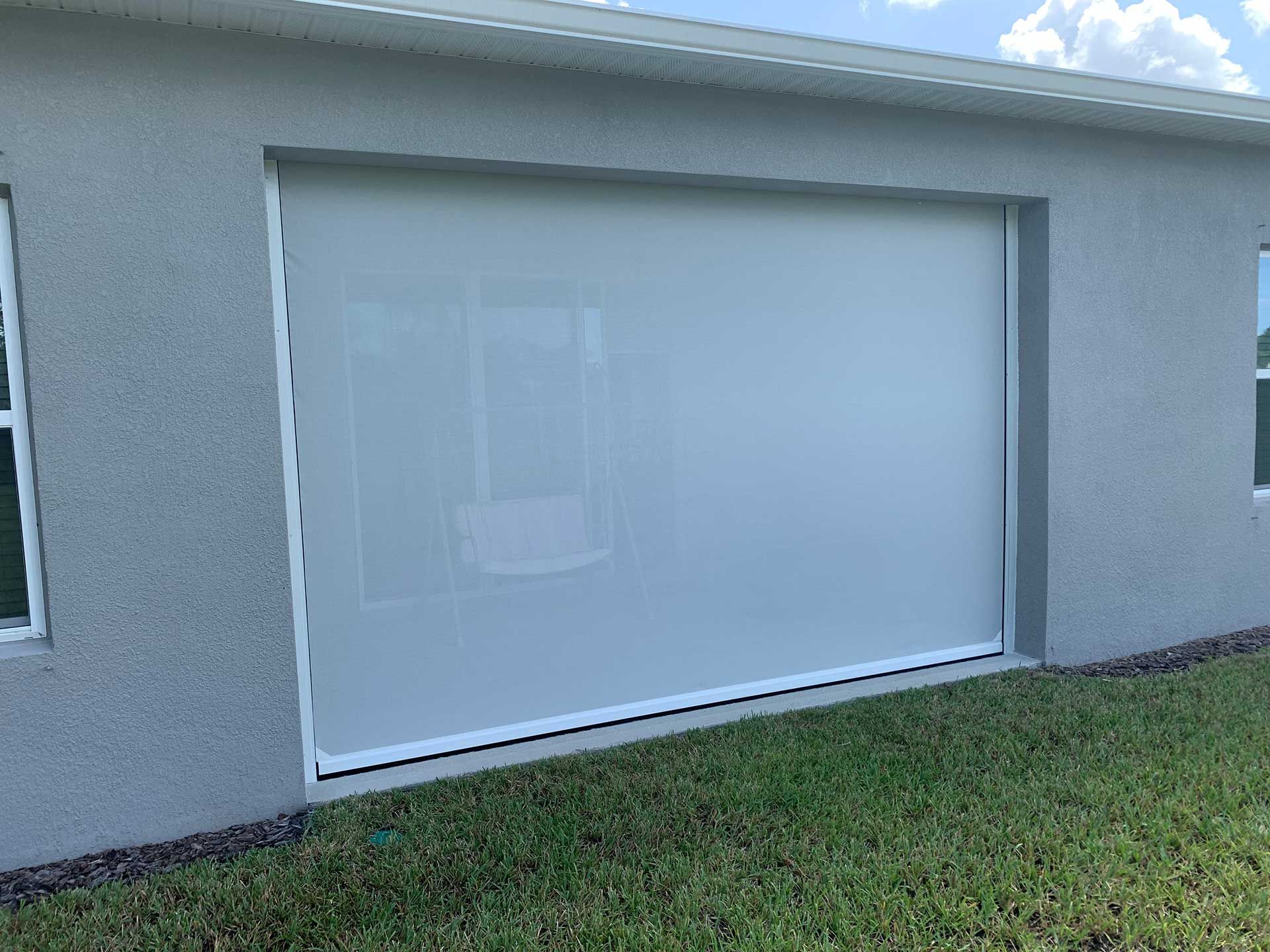 White roller shade covering a large window on a gray house exterior with grass in front