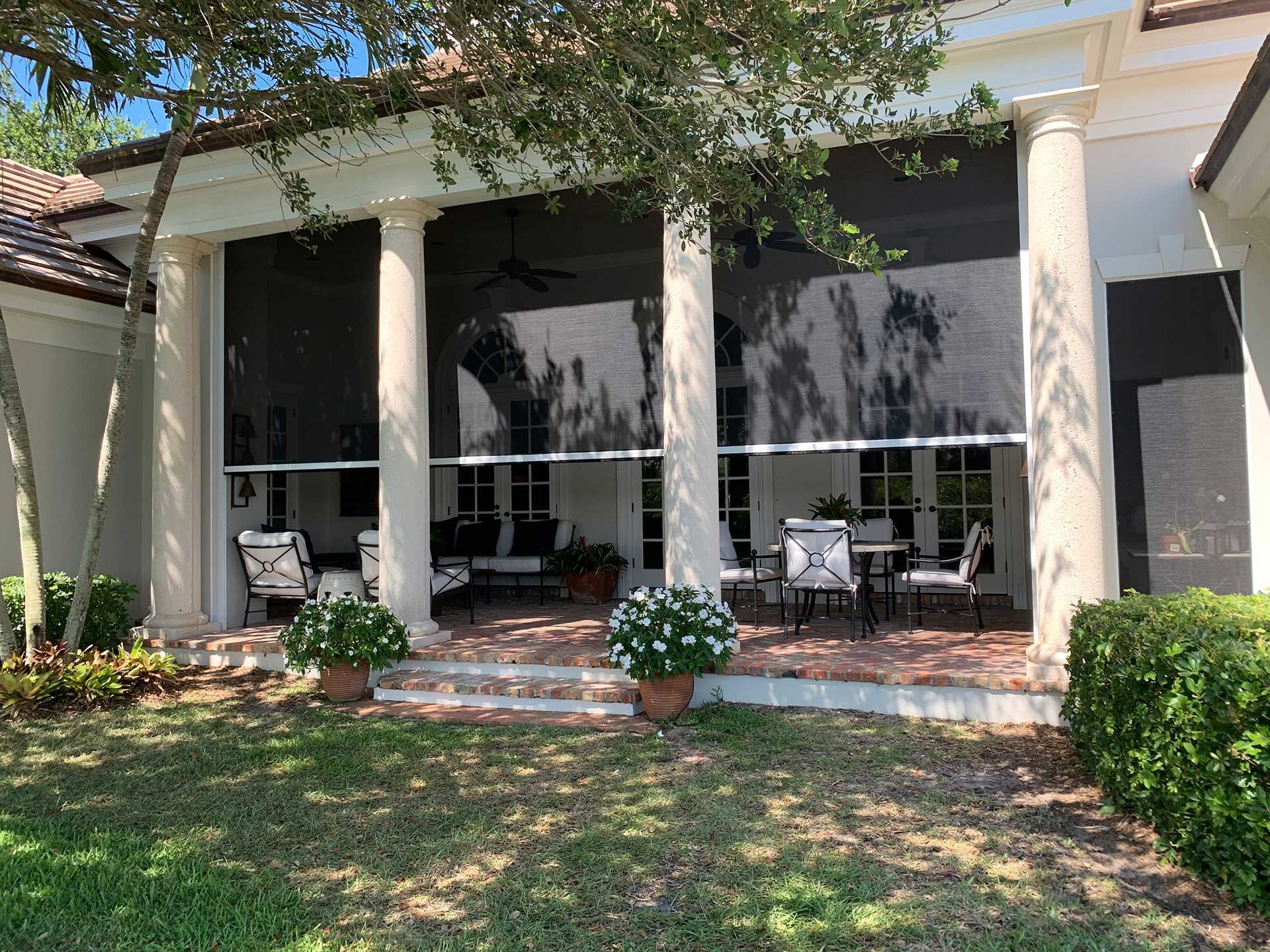 Front porch with white columns, black shutters, and patio chairs under a tree, bordered by shrubs and grass