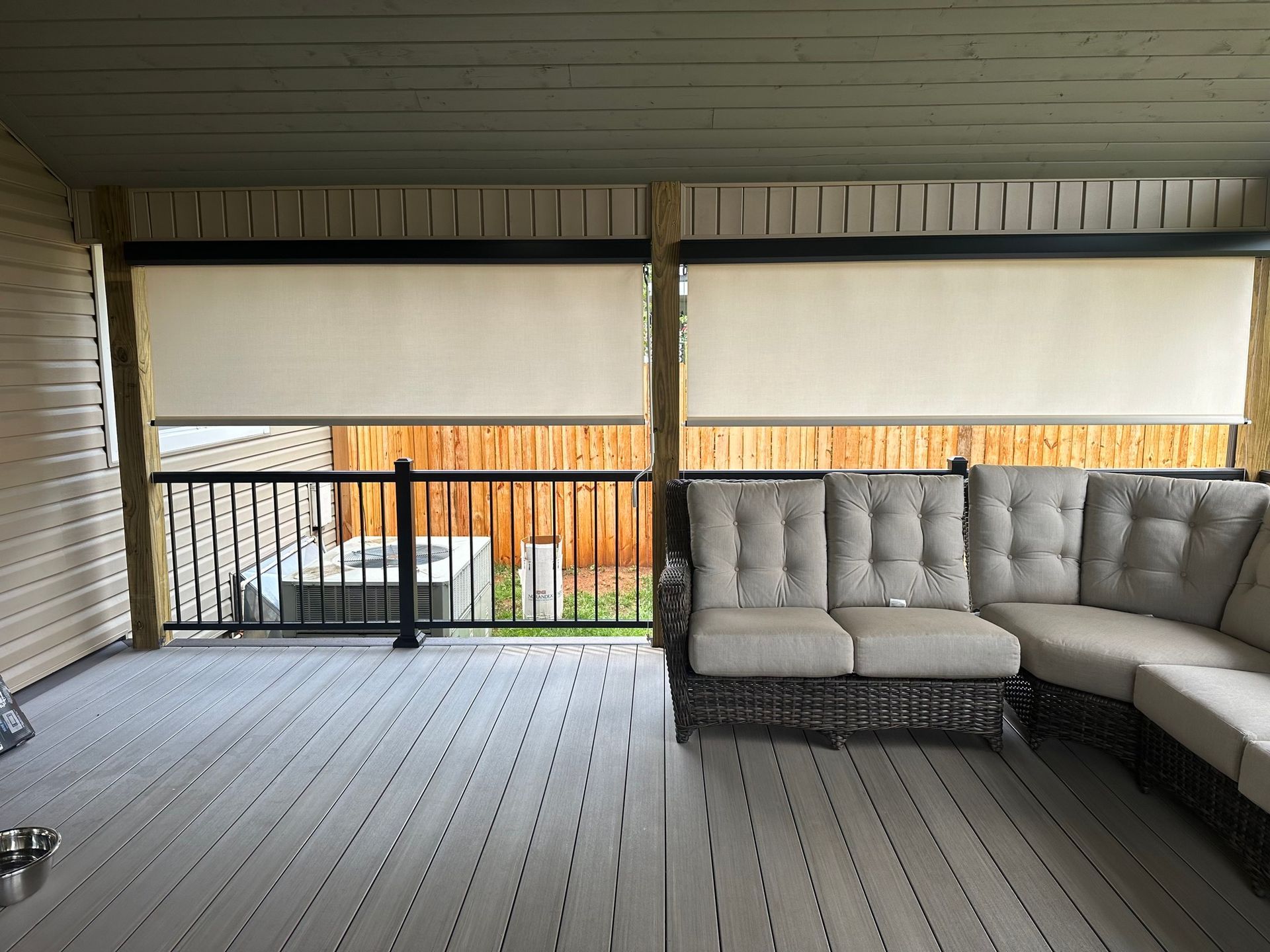 Two beige outdoor roller shades partially down on a wooden deck with seating.