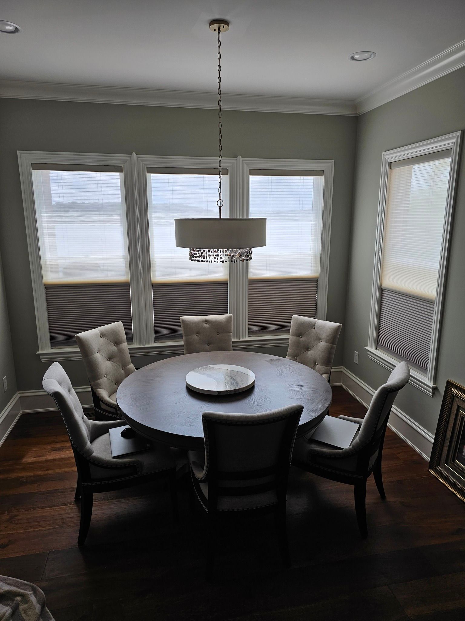 A dining room with a round table, six chairs, and a chandelier. Windows with blinds.
