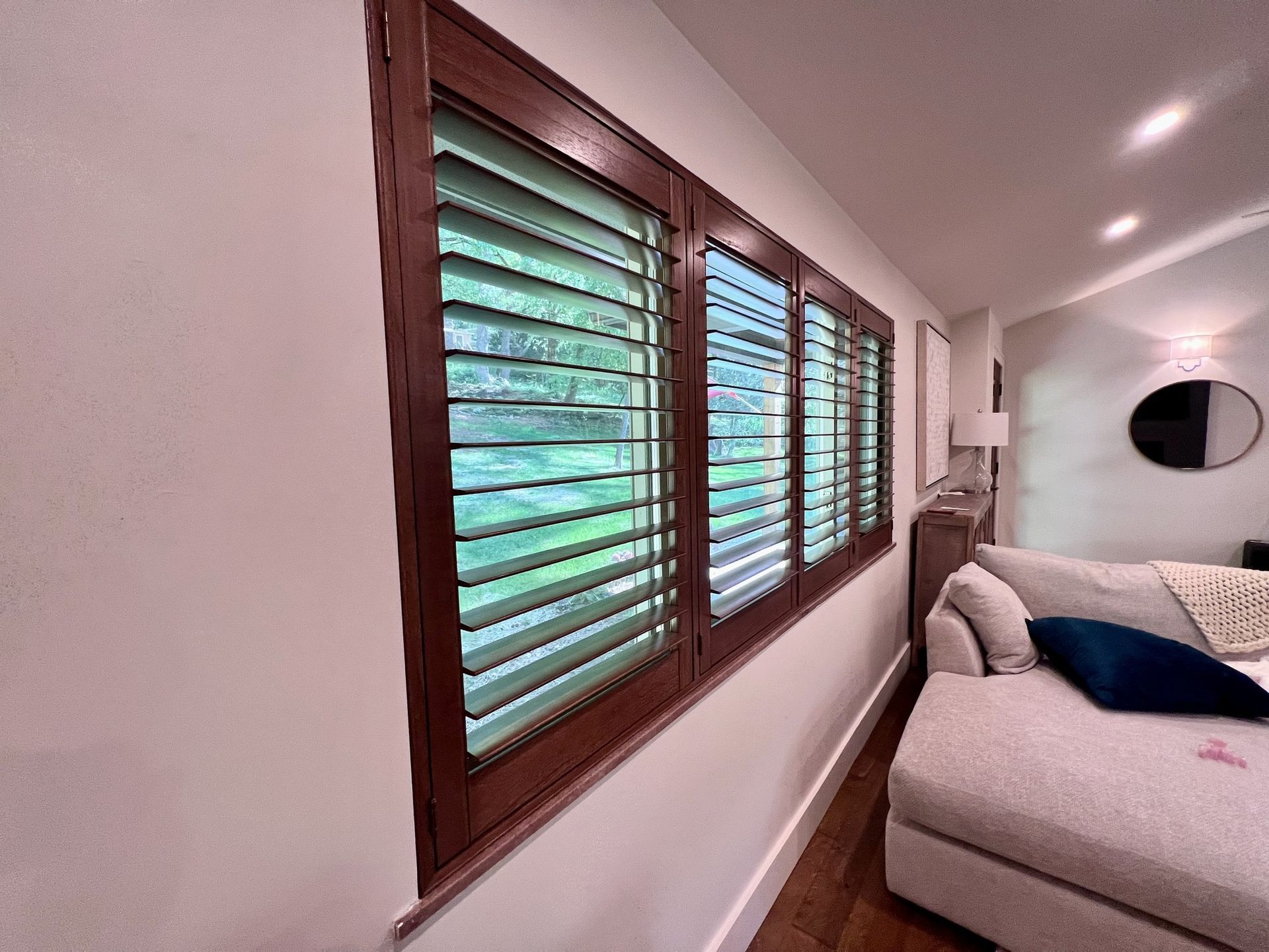 Wooden window shutters on a white wall in a living room, with a couch and round mirror visible.