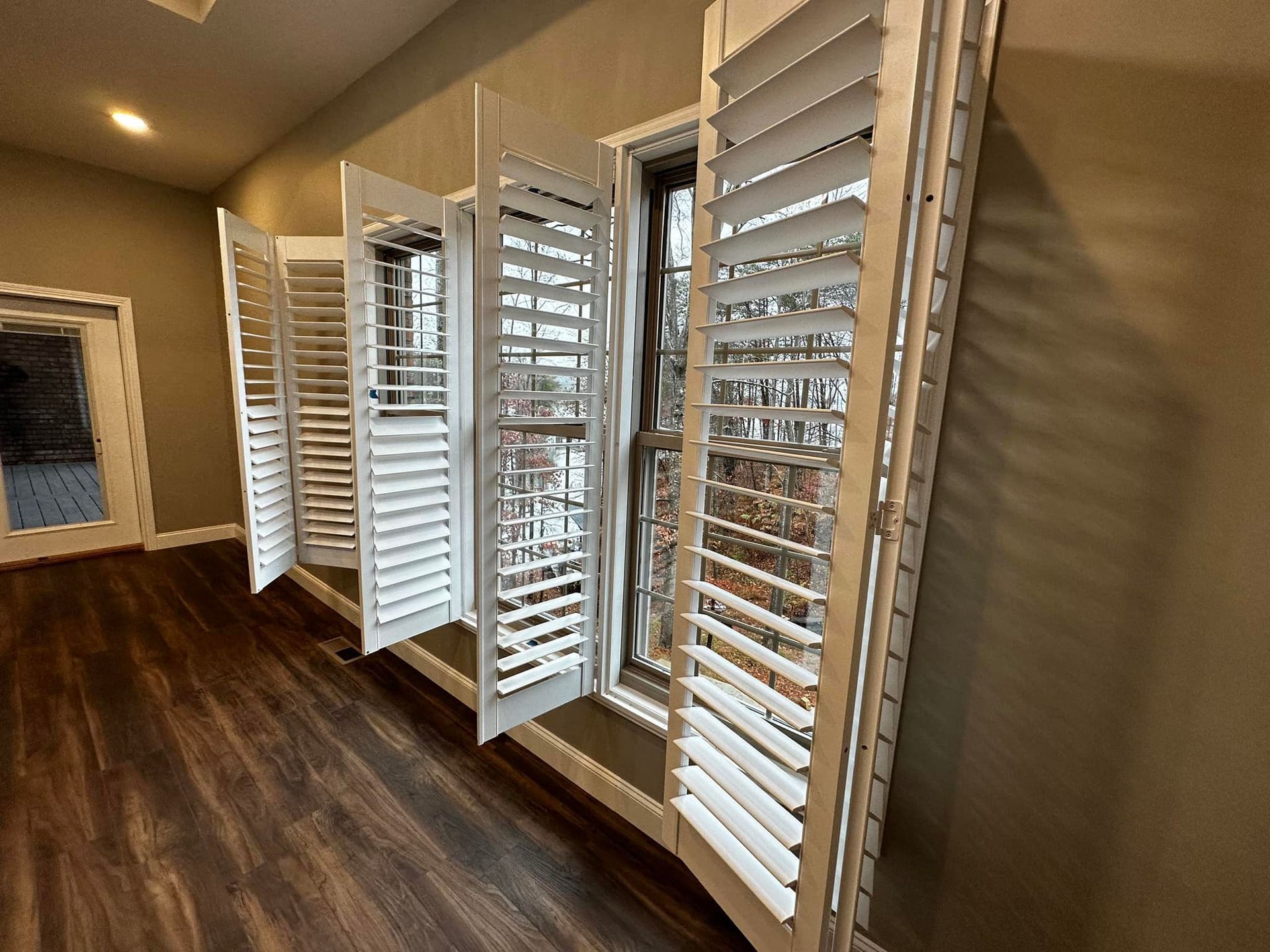 White shutters open on a window in a room with hardwood floors and beige walls.