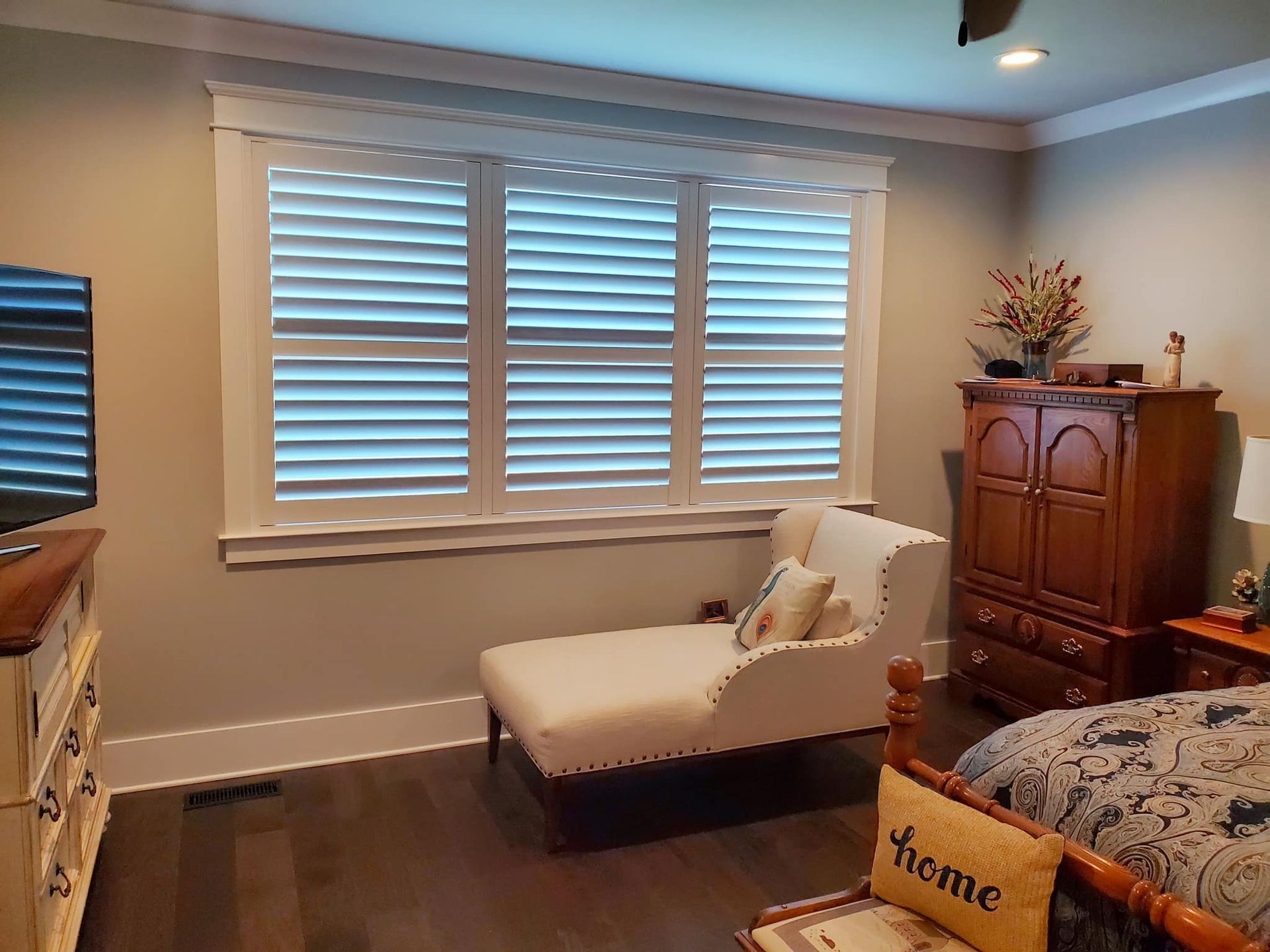 Bedroom with white shutters, chaise lounge, dark wood furniture, and hardwood floors.