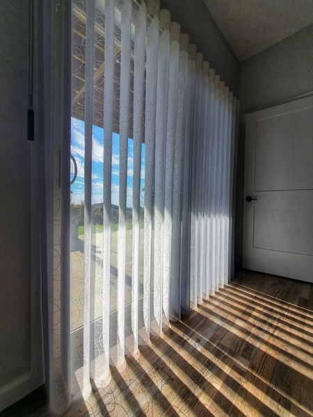 Sheer white vertical blinds filter sunlight through a large window, casting shadows on the wood floor.