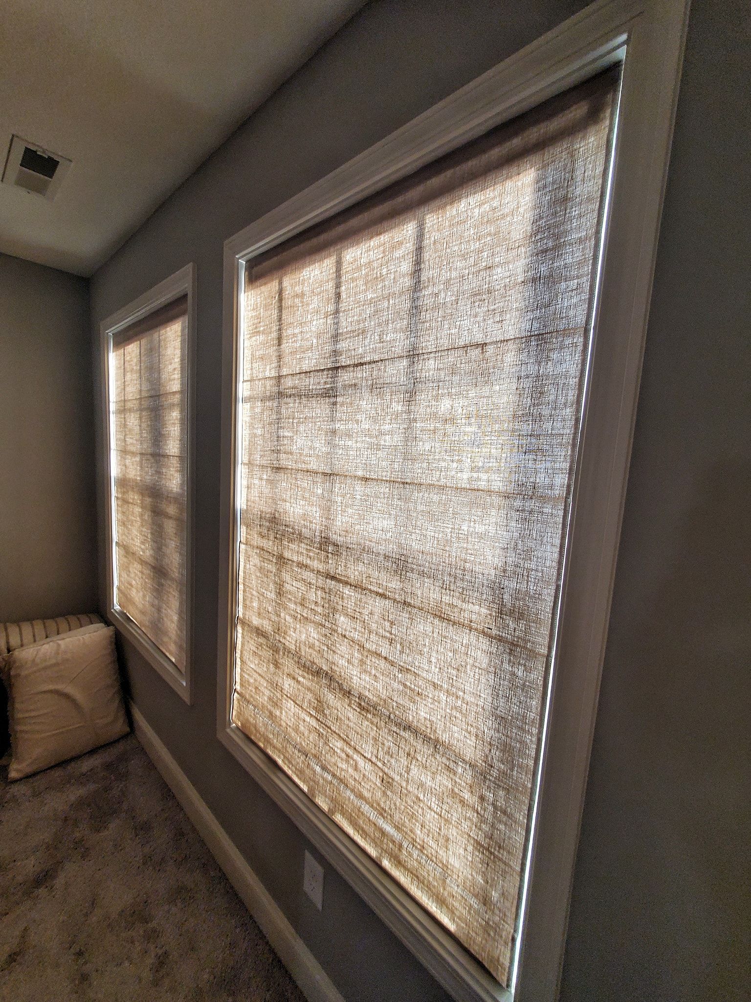 Two windows with woven shades casting shadows; interior wall with a small pillow.