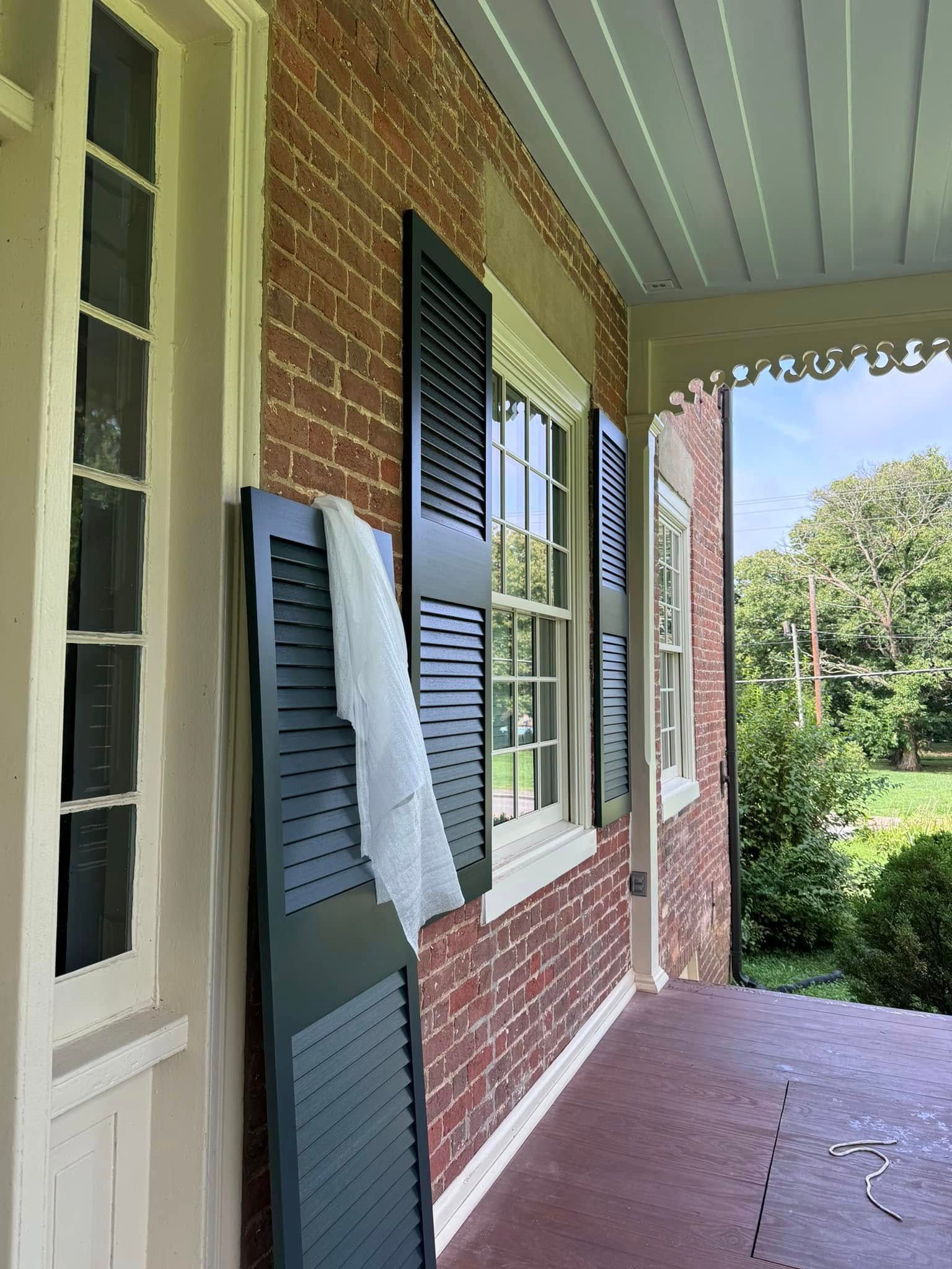 Red brick house exterior with black shutters and white trim, open porch with a white curtain draped on a shutter.