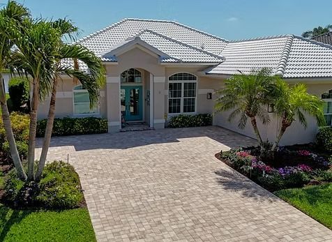 House with tan exterior, turquoise door, and brick driveway, flanked by palm trees and landscaping.