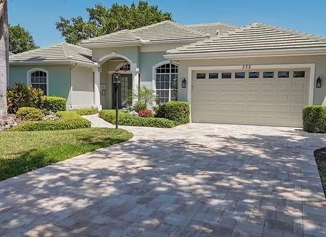 House with light blue exterior, tan garage, and brick driveway. Landscaping and palm tree in yard.
