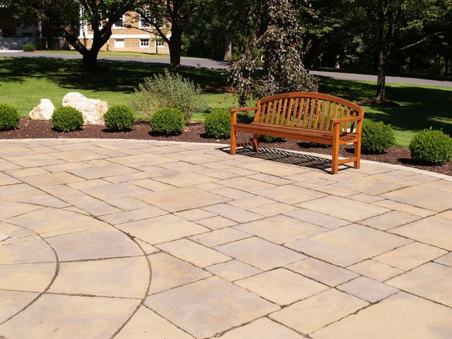 Patio with patterned paving stones, wooden bench, shrubs, and lawn.