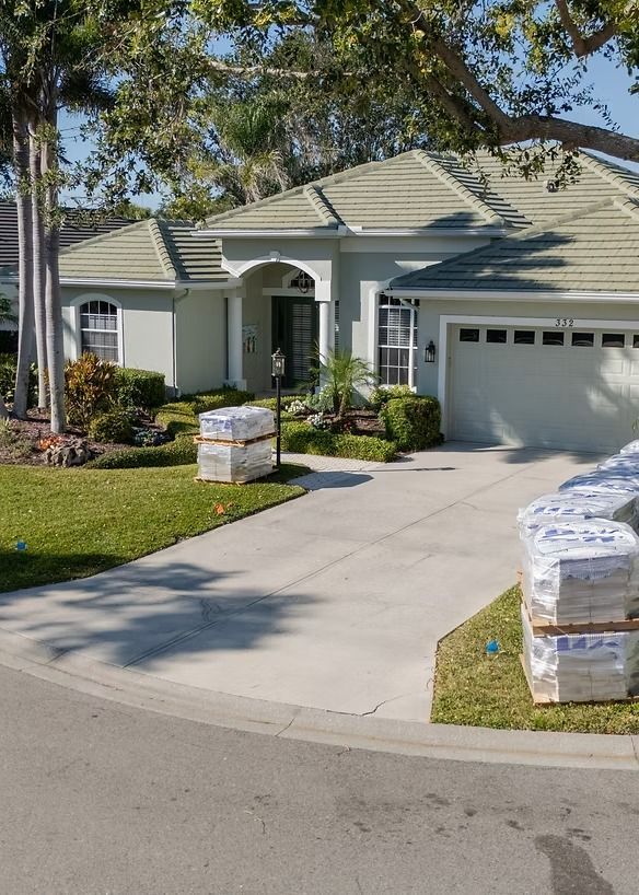 A light blue house with a gray roof and white garage door, with stacks of building materials on the driveway.
