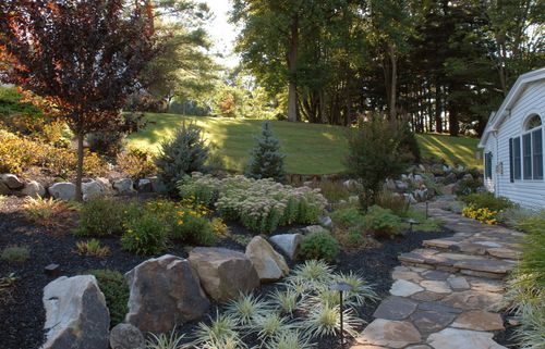 A stone path leads past a landscaped garden with large rocks, shrubs, and a grassy hill towards a white building.