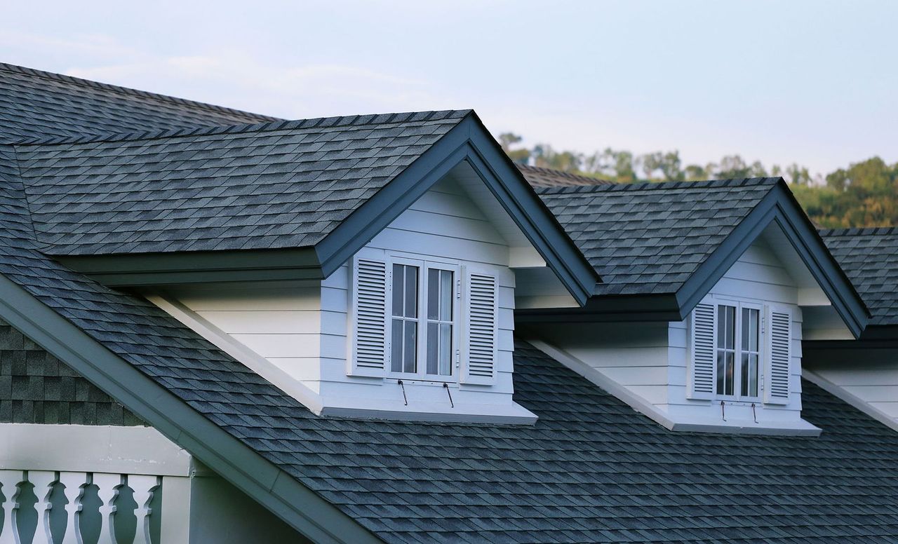 Two dormer windows with white shutters and frames set into a dark gray shingled roof under a light sky.