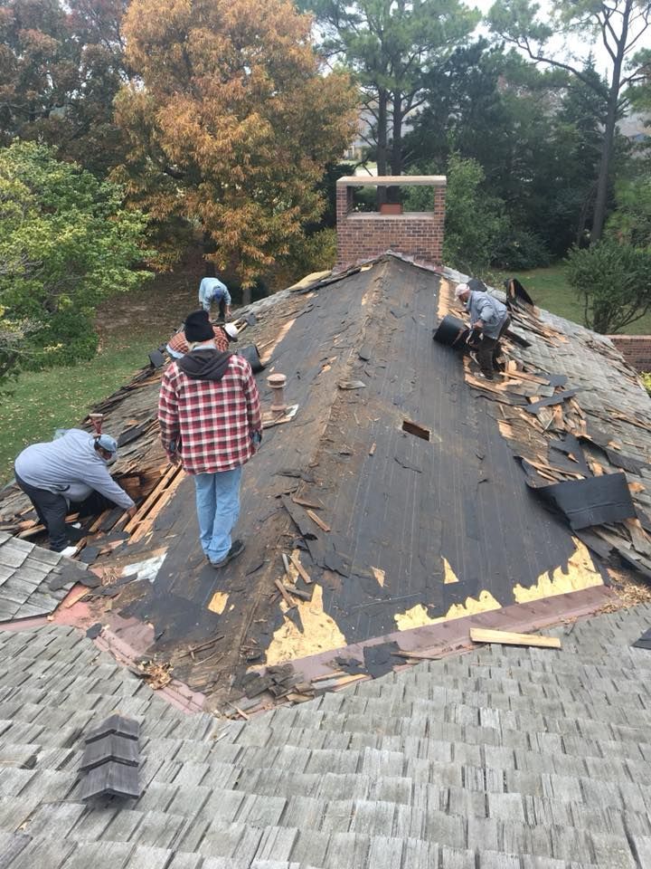 Roofers removing old shingles from a house, standing on a slanted roof next to a chimney.