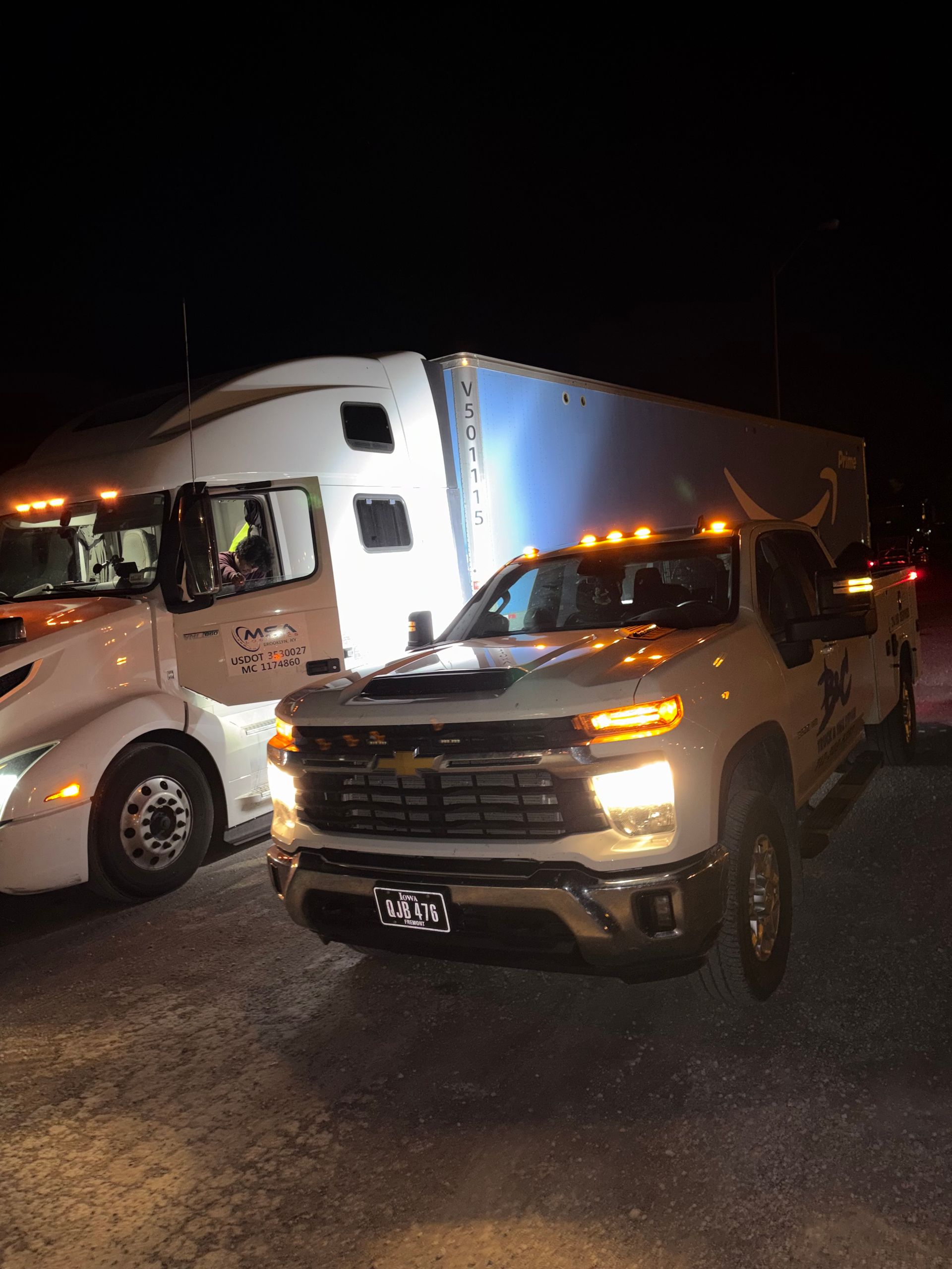 White semi-truck and beige pickup truck parked side by side at night with headlights and amber lights on.