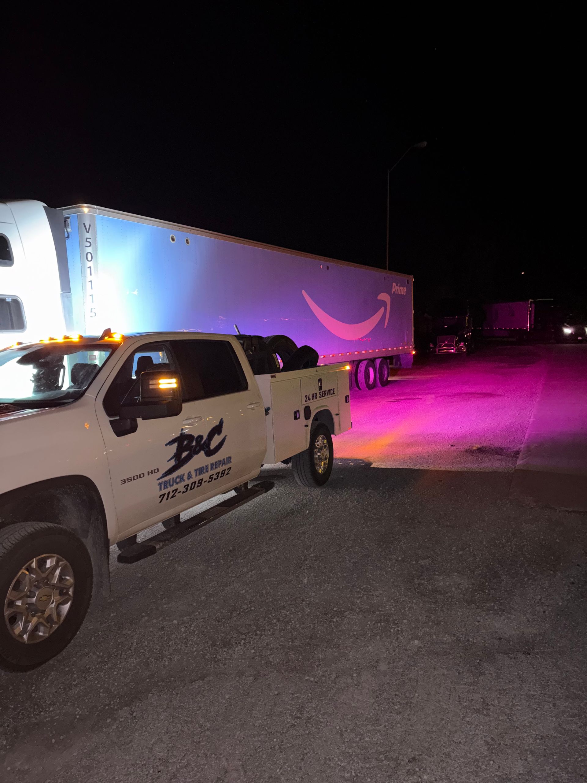 White pickup truck with logo parked beside a lit white trailer at night, washed in purple light.