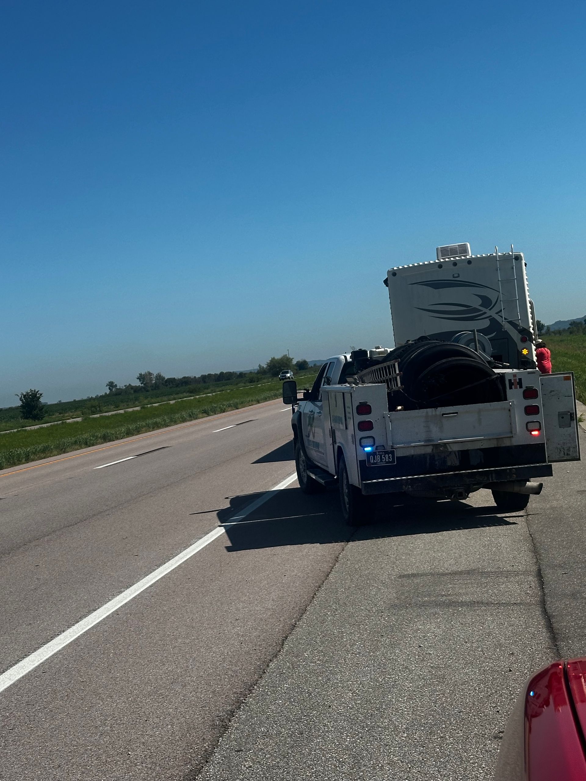 White utility truck with flashing lights stopped on a highway shoulder under a clear blue sky.