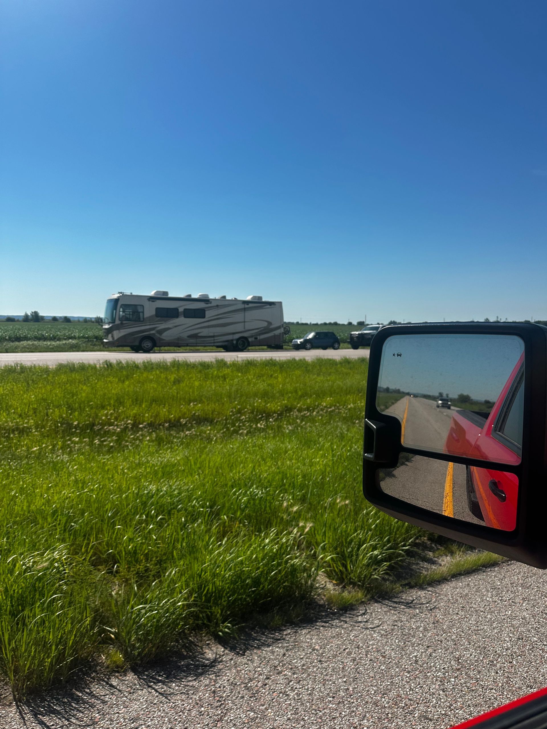 Motorhome driving past a grassy roadside under a clear blue sky, seen beside a car mirror