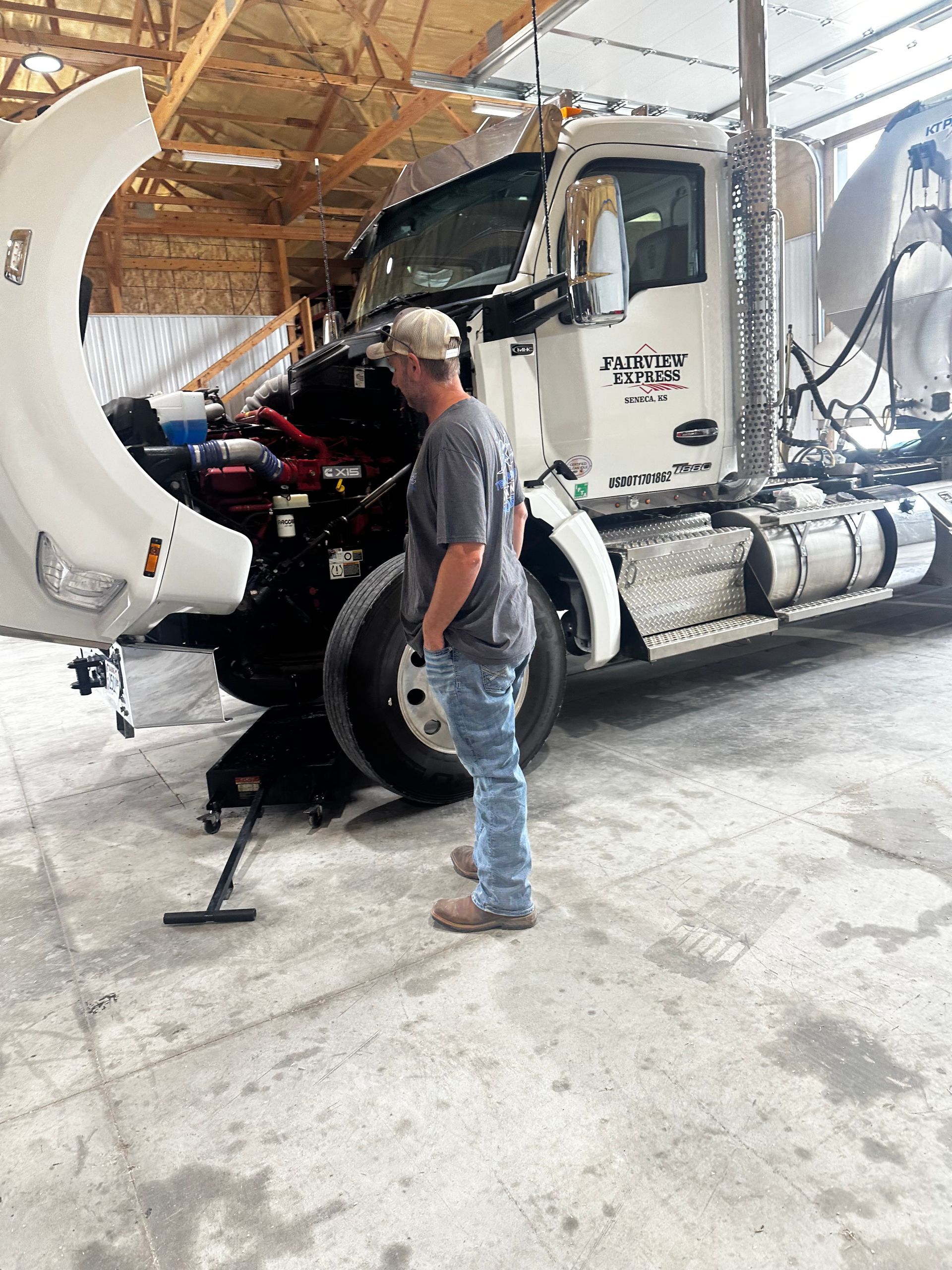Man in gray shirt standing beside a white semi-truck in a garage, looking at the open front end