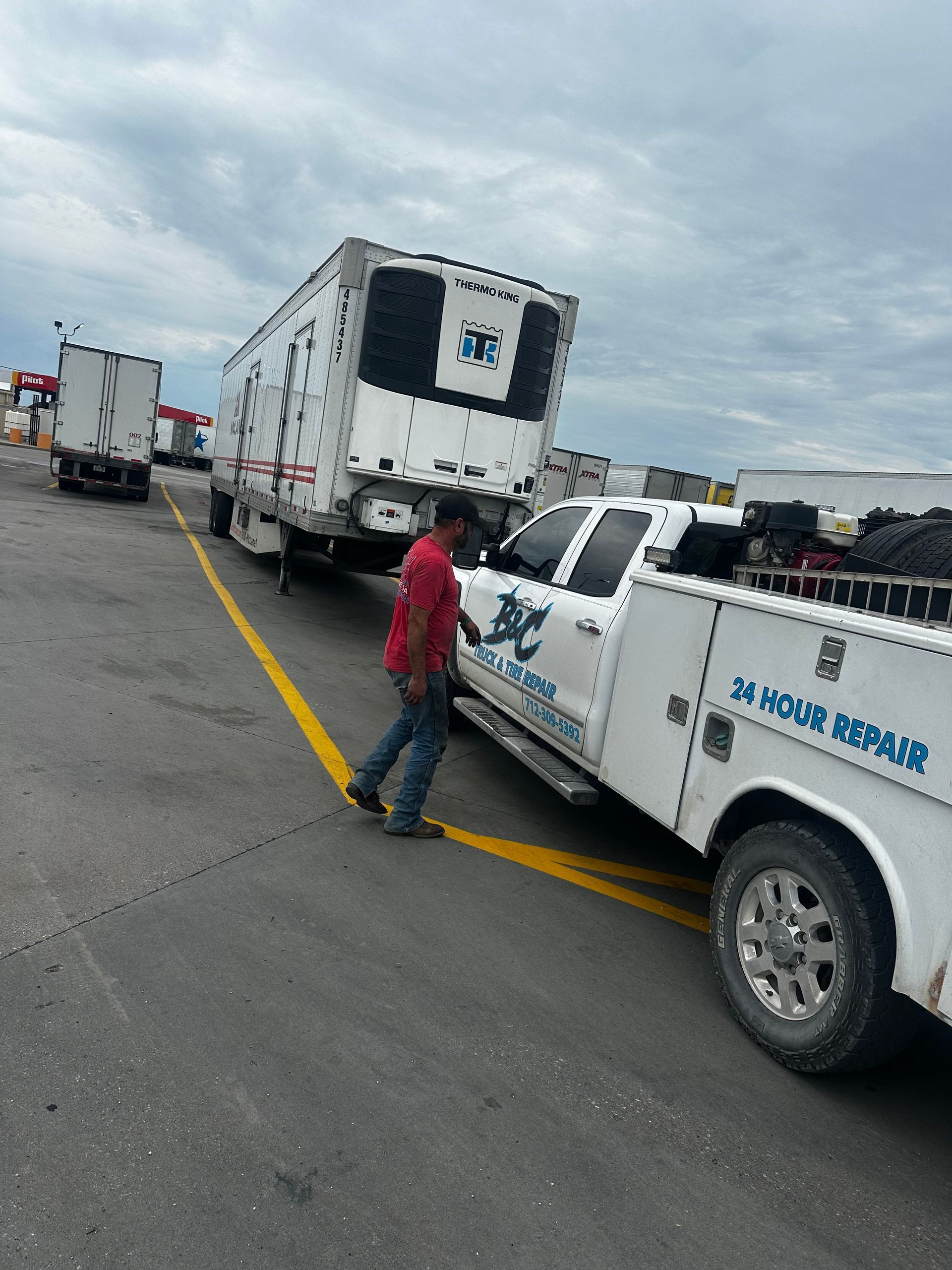 Tow truck with a damaged truck on a highway shoulder, with a worker standing beside it under cloudy skies