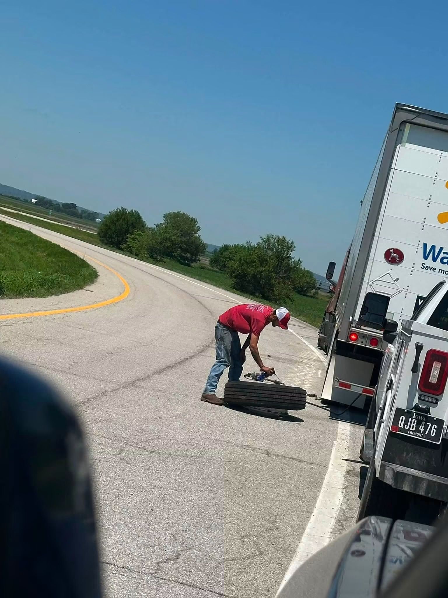 Worker kneeling beside a truck on a highway shoulder, handling equipment near the rear wheel.