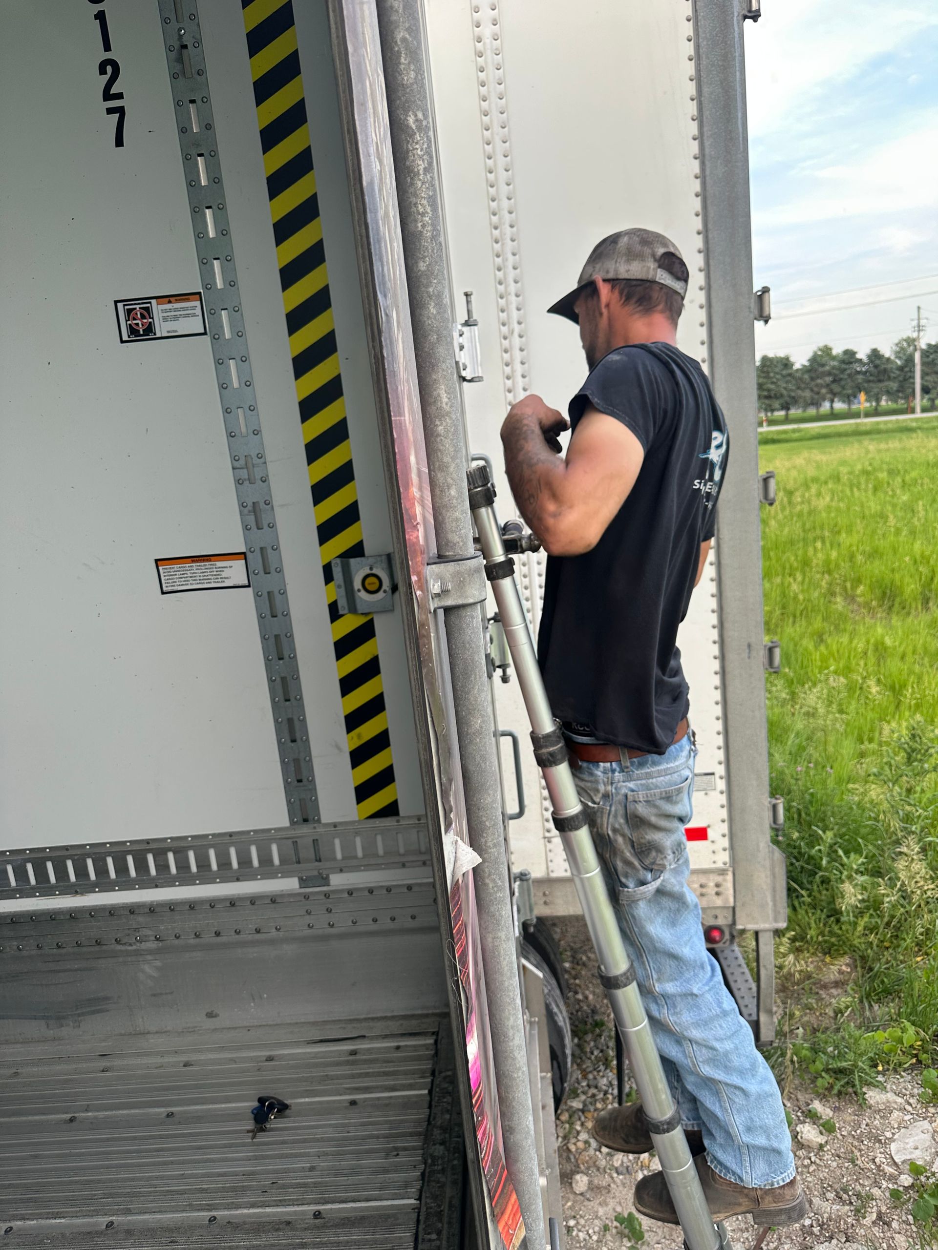 Worker standing on a ladder beside a truck trailer door, adjusting equipment in a grassy roadside area.