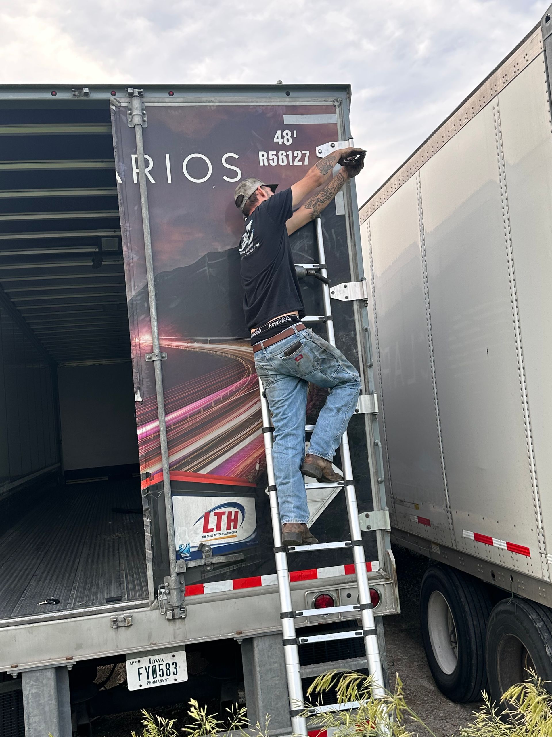 Man climbing the back of a delivery truck using a ladder, with another trailer beside it