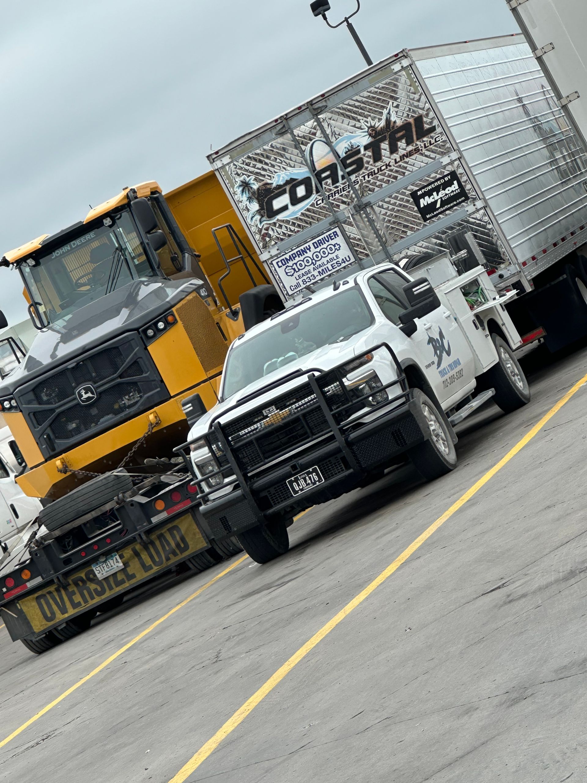 White pickup truck driving on a slanted road beside a yellow semi-truck and silver trailer under cloudy skies