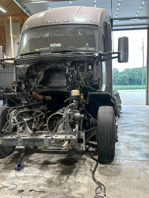 Gray semi-truck cab with front engine removed, parked inside a workshop on a concrete floor.