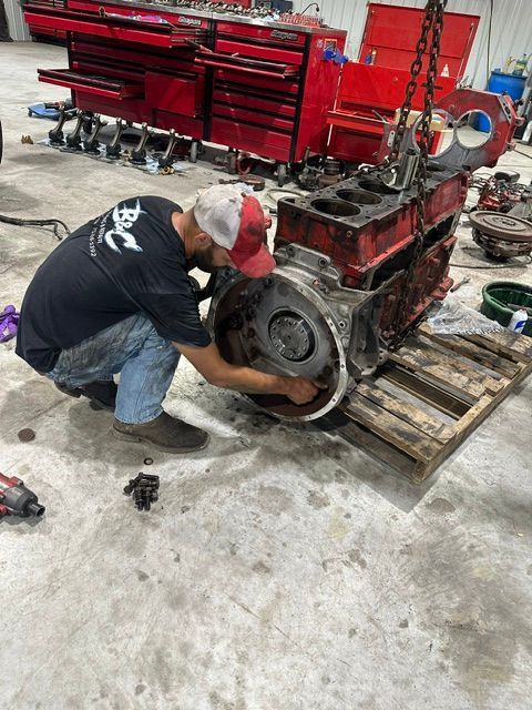 Mechanic kneels beside a red engine on a pallet in a workshop, working with tools.
