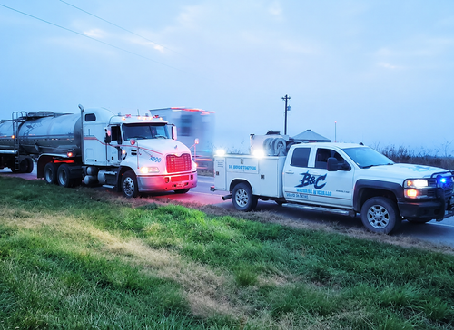 Tow truck and pickup on a roadside at dusk, hauling a tanker truck in rainy conditions