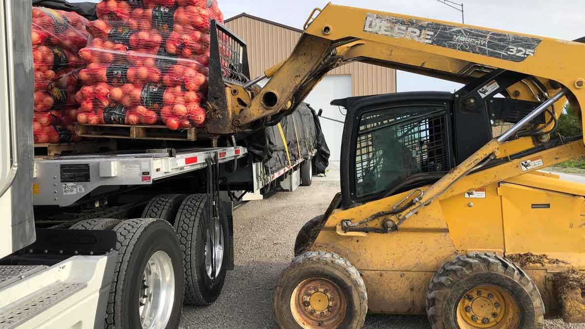 Yellow skid steer unloading red produce crates from a truck bed in an outdoor work yard