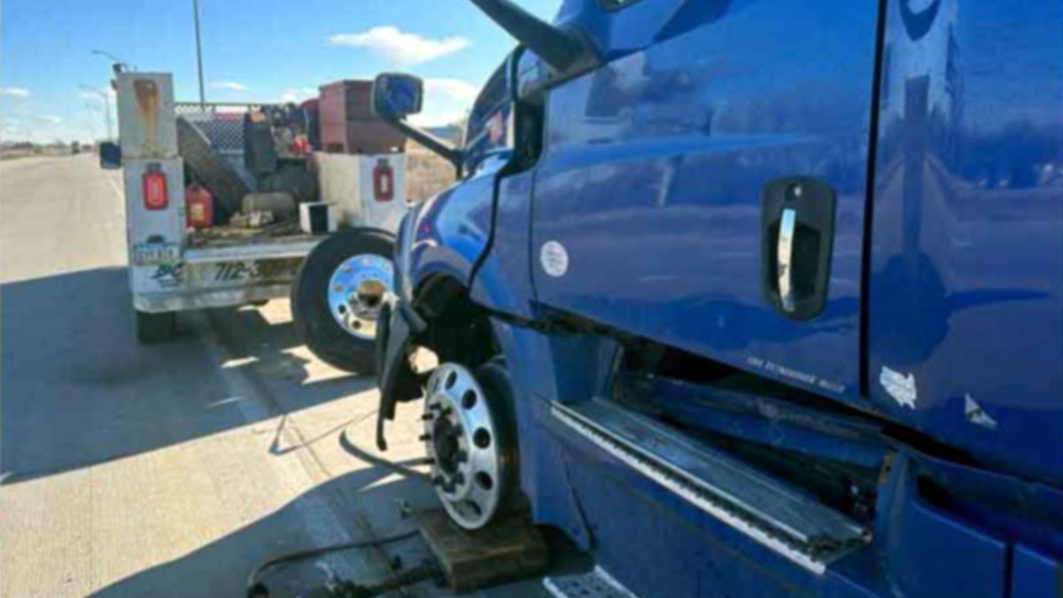Blue semi-truck after a crash, with a damaged front wheel and tow truck behind it on a sunny road.