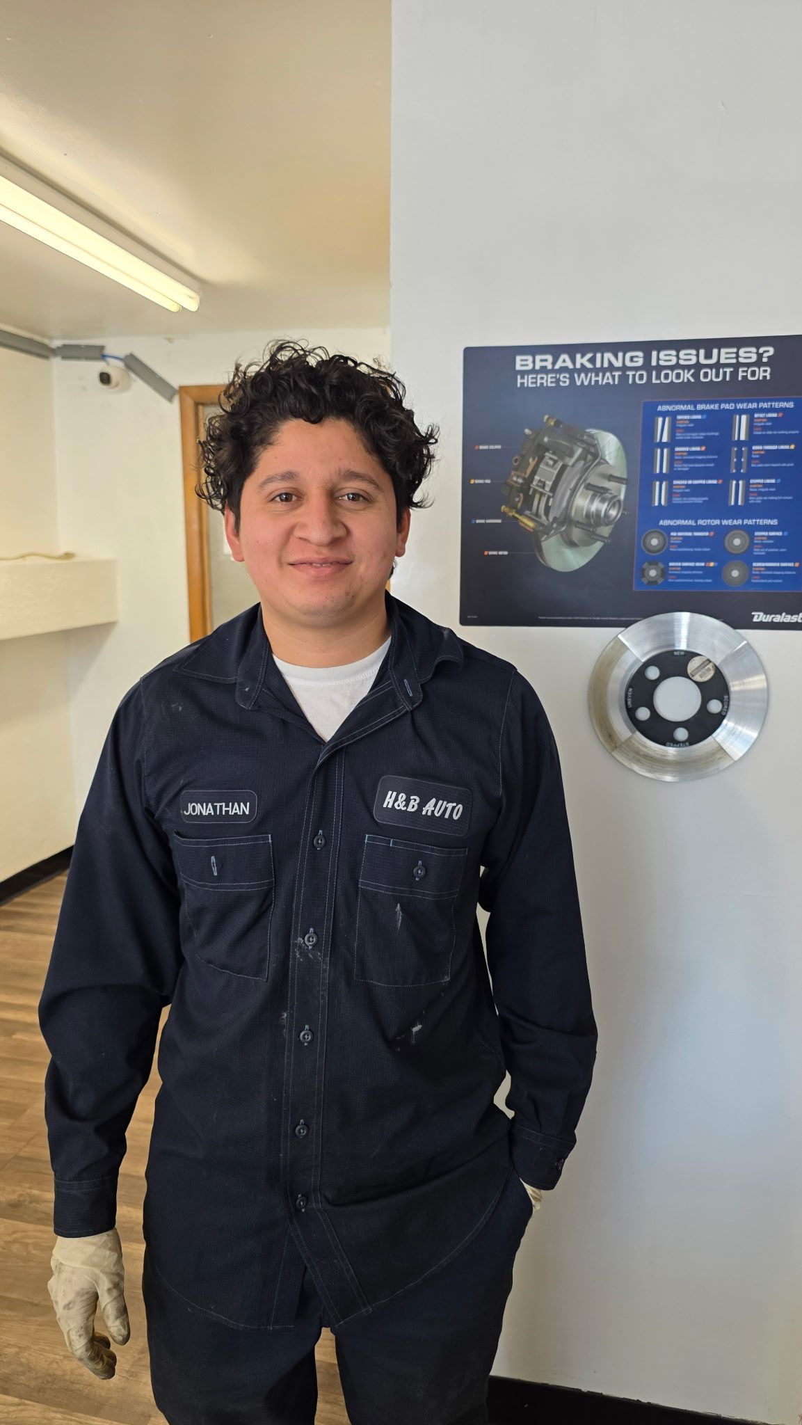 A person wearing a navy polo shirt with an H&B logo stands in an auto repair shop with a car on a lift behind them.