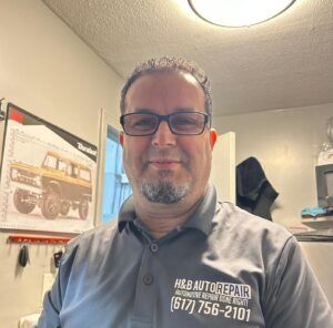 A man in a grey H&B Auto Repair polo shirt stands in an auto repair shop with a vehicle poster on the wall behind him.