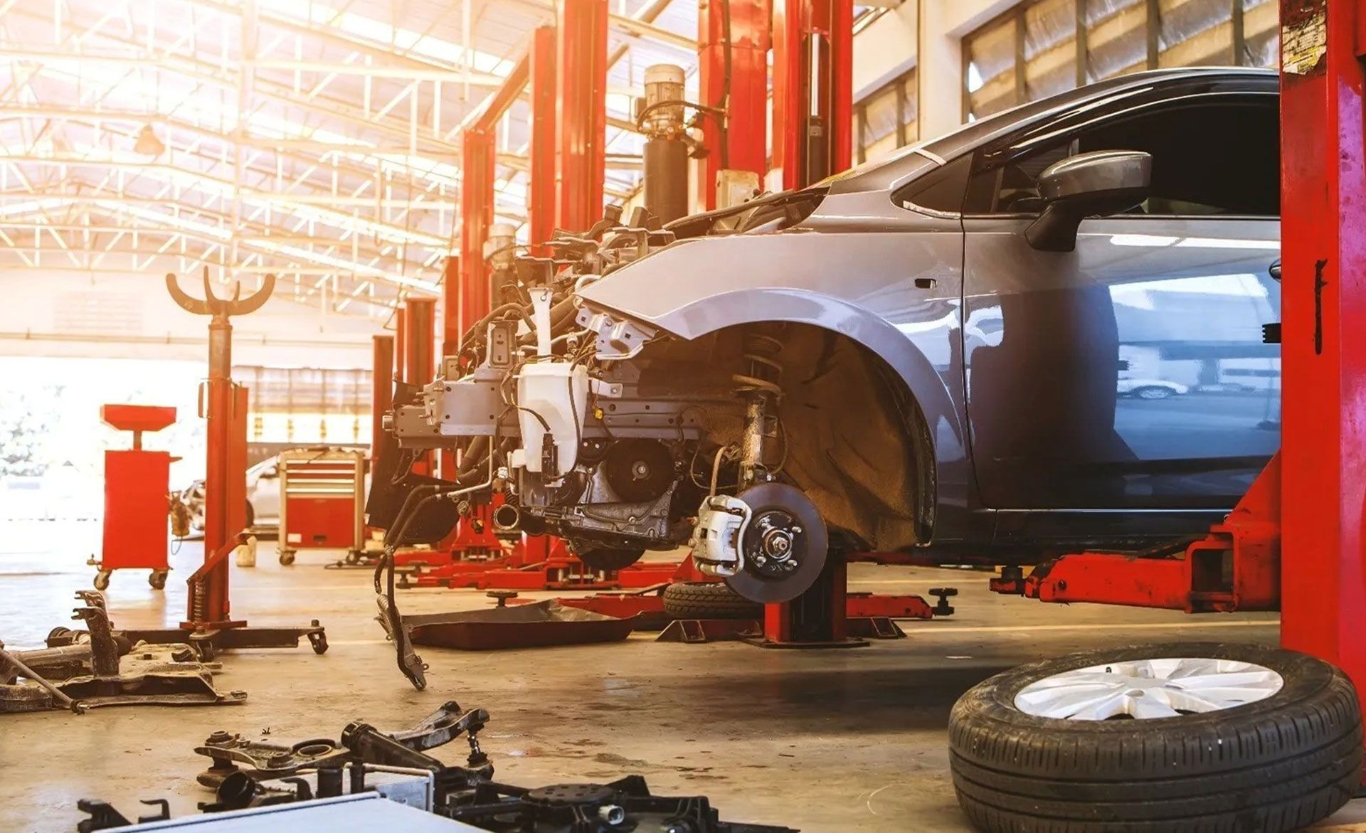 A car undergoing repairs inside a bright, sunlit auto shop, with parts scattered on the floor and a detached tire nearby.